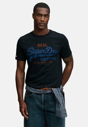Mann trägt ein schwarzes SuperDry-Logo-T-Shirt, blaue Jeans und ein blau kariertes Hemd, das um die Taille gebunden ist, und steht mit neutralem Gesichtsausdruck.