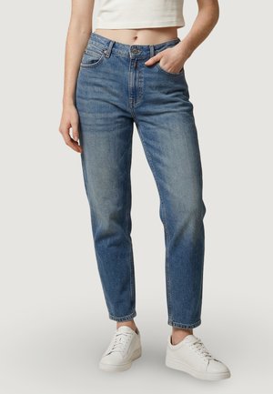 Persona che indossa jeans a vita alta azzurri, top bianco corto e scarpe da ginnastica bianche, in piedi con una mano in tasca contro uno sfondo semplice.