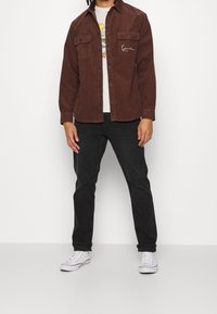 Giacca camicia in corduroy marrone con due tasche sul petto, chiusura a bottoni e t-shirt bianca sotto. Pantaloni neri e sneakers bianche completano l'outfit.