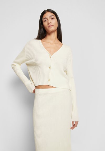 Cardigan canelado de cor creme, com decote em V, fecho de botões e design cropped, combinado com uma saia canelada correspondente.