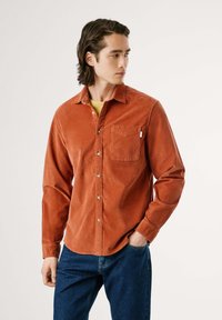 Roodbruine corduroy shirt met knopen, een enkele borstzak en lange mouwen, gedragen met een blauwe spijkerbroek. Soepele textuur en casual pasvorm.