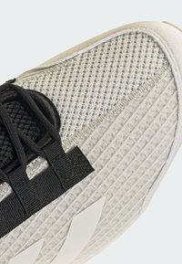 Gros plan de l'embout d'une sneaker blanche avec un tissu en maille texturé, des lacets noirs et un logo angulaire blanc sur le côté.