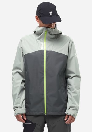 Man wearing two-tone gray Millet jacket with neon green zipper and black Millet cap, looking downward against plain background.