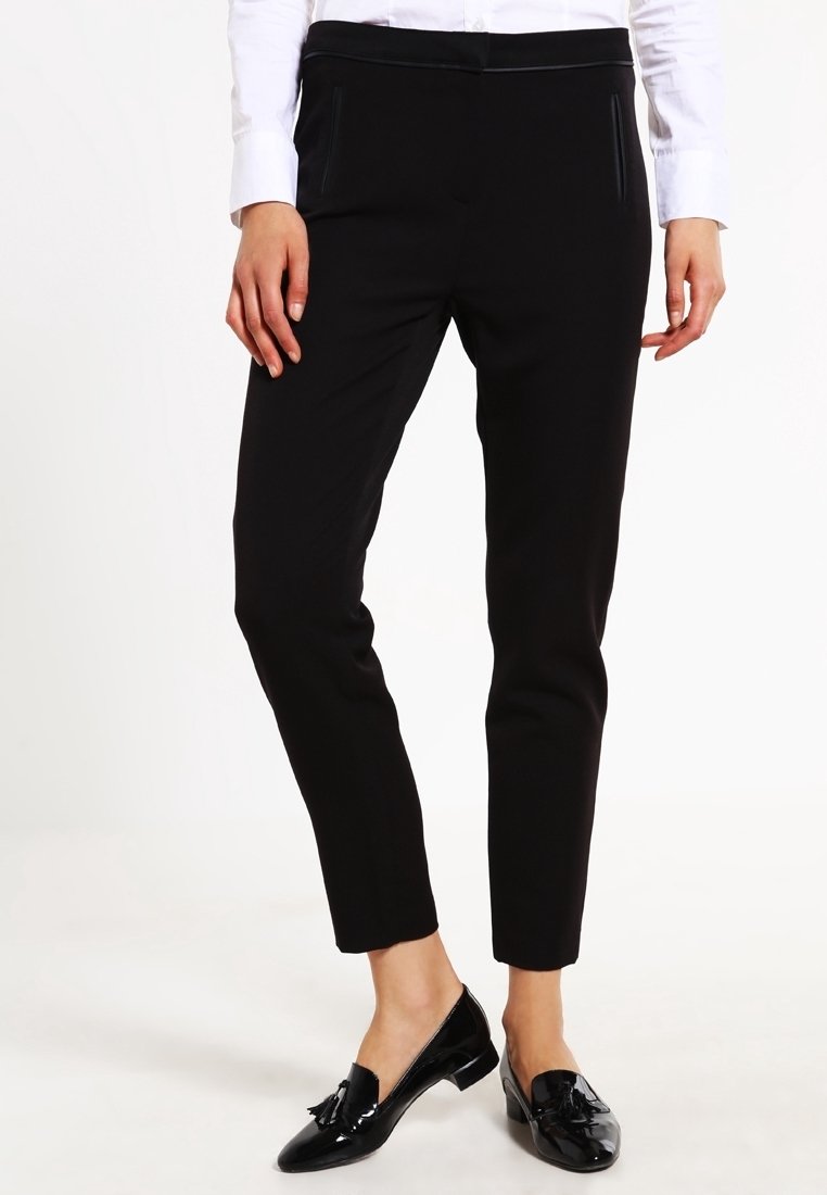 Femme portant un pantalon noir slim, une chemise blanche à manches longues et des mocassins noirs brillants avec des glands, debout devant un fond blanc uni.