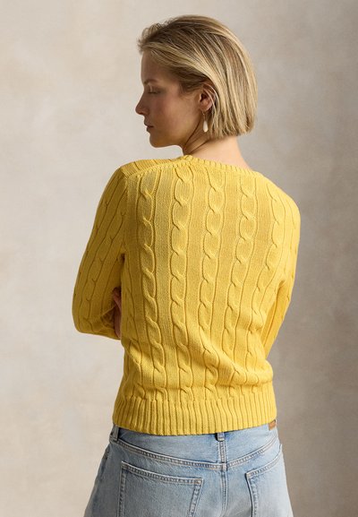 Γυναίκα με κοντά ξανθά μαλλιά, φορώντας κίτρινο πουλόβερ πλεκτό με πλεξούδες και ανοιχτό μπλε τζιν, στέκεται μπροστά σε ουδέτερο φόντο.