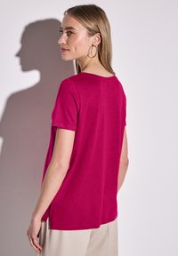 Haut fuchsia à manches courtes avec col rond, coupe décontractée, fentes latérales et texture lisse, porté avec un pantalon de couleur claire.