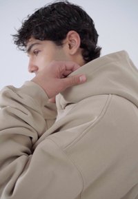 Sweat à capuche beige en tissu doux et texturé, doté d'une grande capuche et de poignets côtelés, présenté en train d'être ajusté au niveau du col.