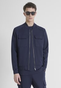 Chaqueta navy con cremallera, dos bolsillos en el pecho, puños acanalados y una textura suave, llevada sobre una parte superior y pantalones a juego, combinada con gafas de sol.
