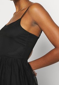 Kvinna iklädd en ärmlös svart klänning med tunna remmar och lager på lager kjol, som visar sidan och armen mot en vit bakgrund.