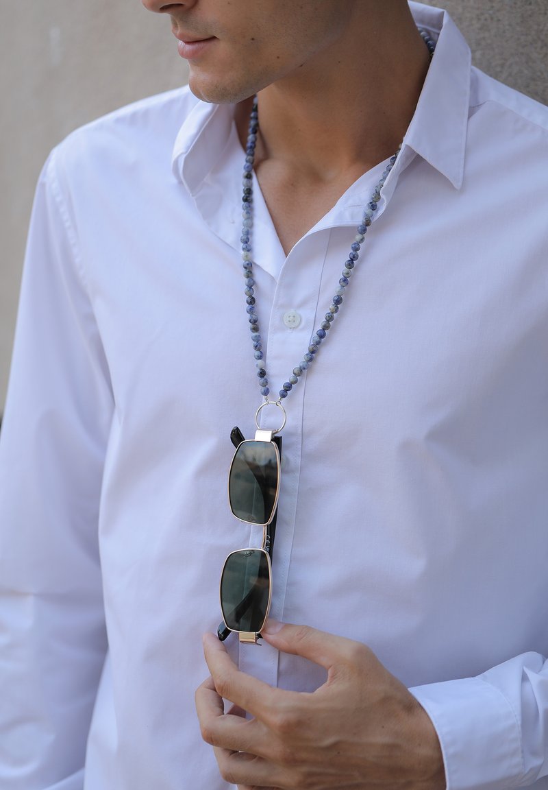 De grandes lunettes de soleil noires avec des accents dorés pendent d'une chaîne en perles bleues. La personne porte une chemise blanche à boutons.