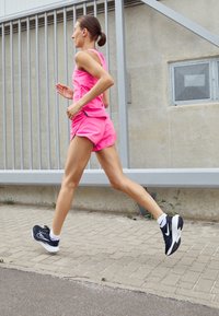 Coureur dans un débardeur rose vif et un short, portant des chaussures de sport noir et blanc, sur une surface pavée texturée.