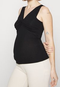 Svart ärmlös topp med V-ringning och rynkad framdesign, gjord av mjukt tyg. Matchad med ljusa ribbade byxor.