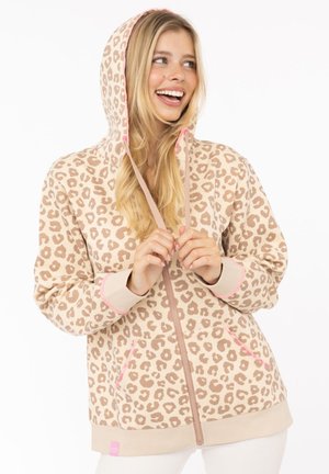 Lächelnde Frau, die einen beige-pinken Leopardenprint-Zip-Hoodie trägt, hält die Kapuzenkordeln und schaut seitlich vor einem weißen Hintergrund.