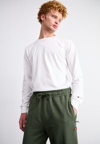 Camisa blanca de manga larga combinada con pantalones de chándal verdes con cordón, que presentan bolsillos laterales y un pequeño logotipo en el muslo izquierdo.