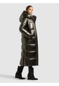 Garš, pūkains parka mētelis no spīdīga tumši zaļa auduma ar kapuci, ar šūtiem laukumiem un taisnu griezumu, kopā ar melnām platformas zābakiem.