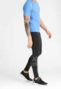 Svarta träningsbyxor med grå ränder, i kombination med en kortärmad blå skjorta och svarta sneakers, med en slät textur och en kroppsnära design.