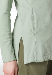 Långärmad topp i ljusgrön bomull, med sidoklipp och synliga sömsdetaljer, ihopparad med olivgröna byxor.