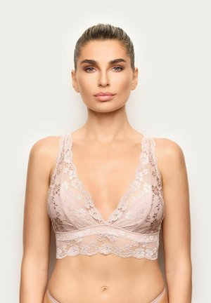 Mujer con el pelo recogido, vistiendo un bralette de encaje rosa claro, mirando al frente sobre un fondo neutro y liso.