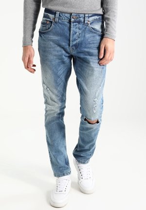 Homme portant un jean bleu clair usé avec une déchirure au genou droit, un t-shirt gris à manches longues et des baskets blanches, marchant vers l'avant.