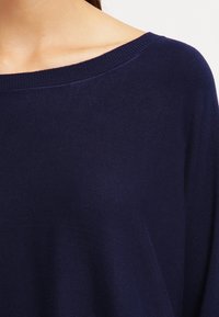 Námořnický modrý svetr s širokým výstřihem a žebrovaným lemem, vyrobený z měkké, texturované pleteniny. Design má volný střih.