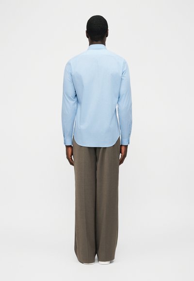 Άνδρας που στέκεται με την πλάτη προς το μέρος μας, φορώντας γαλάζιο πουκάμισο με μακριά μανίκια και άνετο καφέ παντελόνι, σε απλό λευκό φόντο.