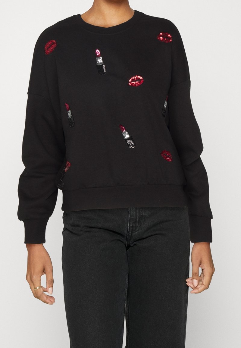 Sweat-shirt noir avec des lèvres en sequins rouges et motifs de rouge à lèvres, porté avec un jean noir, montrant le haut du corps et les mains.