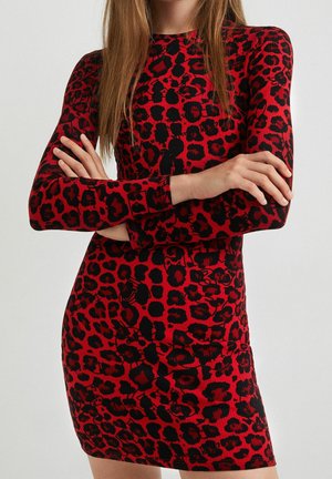 Femme aux longs cheveux portant une robe rouge ajustée avec imprimé léopard noir, debout, les bras croisés devant un fond uni.