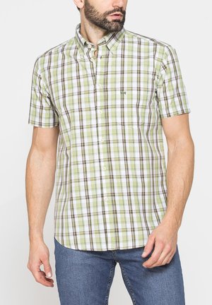 Uomo con camicia a maniche corte a quadri verdi, bianchi e marroni, abbinata a jeans blu, in piedi davanti a uno sfondo semplice.