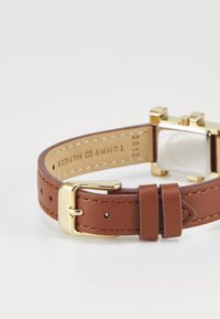 Brown leather watch strap with gold hardware, featuring a square casing and buckle, and visible stitching. Engraved logo on the inner band.
