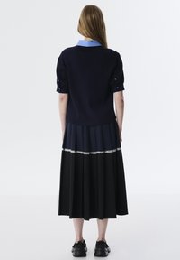 Maglione navy a maniche corte con colletto azzurro, abbinato a una gonna nera plissè con un accento di pizzo bianco. Scarpe nere visibili.