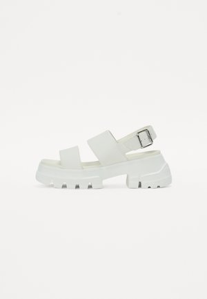 Hvite sandaler med en klumpete såle, med to brede stropper og en spenne detalj på ankelstroppen. Glatt tekstur, minimalistisk design.