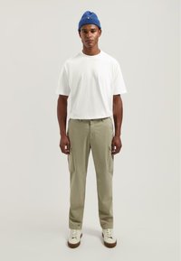 Bílý tričko s krátkým rukávem, světlé zelené cargo kalhoty s bočními kapsami a bílé boty s hnědými podrážkami. Modrá pletená čepice přidává kontrast.