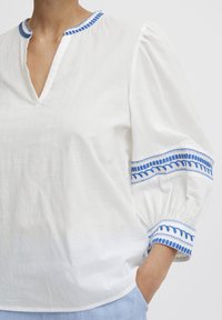 Blouse blanche avec un décolleté en V, accents brodés bleus sur le col et les manches, et manches bouffantes. Fabriquée en tissu léger, coupe décontractée.