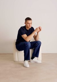 Scaun moale tapițat alb, cu marginile rotunjite, persoană așezată, îmbrăcată cu un tricou bleumarin și blugi, teniși albi, mâinile împreunate în poală.