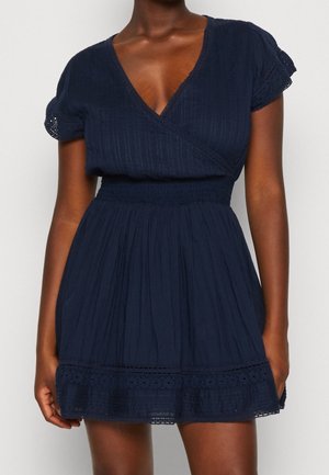 Granatowa sukienka z krótkim rękawem i dekoltem w serek, z marszczonym pasem oraz ażurową koronką na rękawach i dole, noszona przez osobę o ciemnej karnacji.