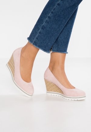 Ben iført korte blå jeans og lyserøde kilehælte sko med flettet tekstur på sålen, stående på hvidt gulv.