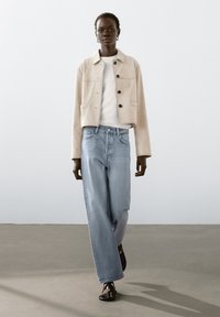 Beige mockasinfodrad jacka med krage och svarta knappar, kombinerad med ljusblå vida jeans. Modellen bär en vit t-shirt och svarta sandaler.