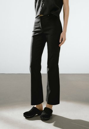 Personne portant un pantalon noir droit et des sabots noirs cloutés, debout sur un sol gris devant un fond blanc.