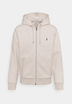Beiger Kapuzenpullover mit Reißverschluss, Fronttaschen, Kordelzug an der Kapuze, gerippten Bündchen und kleinem braunem Logo auf der linken Brust.