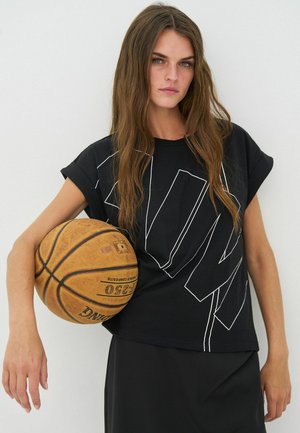 Sort T-shirt med korte ærmer og hvide geometriske linjer, kombineret med et sort nederdel. Modellen holder en slidt basketball mod armen.