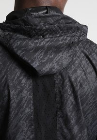 Cortavientos negro con patrón texturizado, que incluye capucha y panel lateral perforado para mayor transpirabilidad. Acabado suave y tejido ligero.