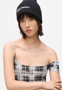 Ung kvinde iført en sort strikbeanie med hvid tekst og en sort-grå plaid off-shoulder top, arme korslagte, neutral baggrund.