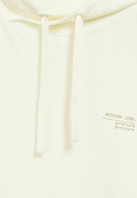 Světle žlutá mikina s kapucí na stahování. Obsahuje potisk s textem "NIC MÉNĚ." spolu s koordináty na straně. Hladká textura.