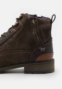 Bottines en suède marron avec des accents en cuir foncé, des éléments en métal, des coutures contrastées et une semelle en caoutchouc texturé. Logo sur le talon.