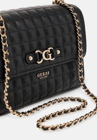 Bolso acolchado negro con una correa de cadena dorada. Presenta un cierre con logo dorado y un logotipo "GUESS" con "EST 1981" grabado en un panel negro.
