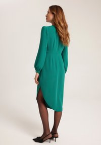 Zielona sukienka w odcieniu emerald z długimi rękawami, dopasowaną sylwetką oraz rozcięciem do uda z tyłu, w połączeniu z czarnymi, spiczastymi szpilkami i przezroczystymi rajstopami.