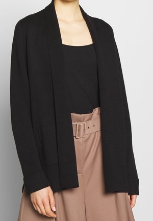 Cardigan aperto nero con vestibilità rilassata, maniche lunghe e tasche laterali, indossato sopra un top nero e pantaloni beige a vita alta.