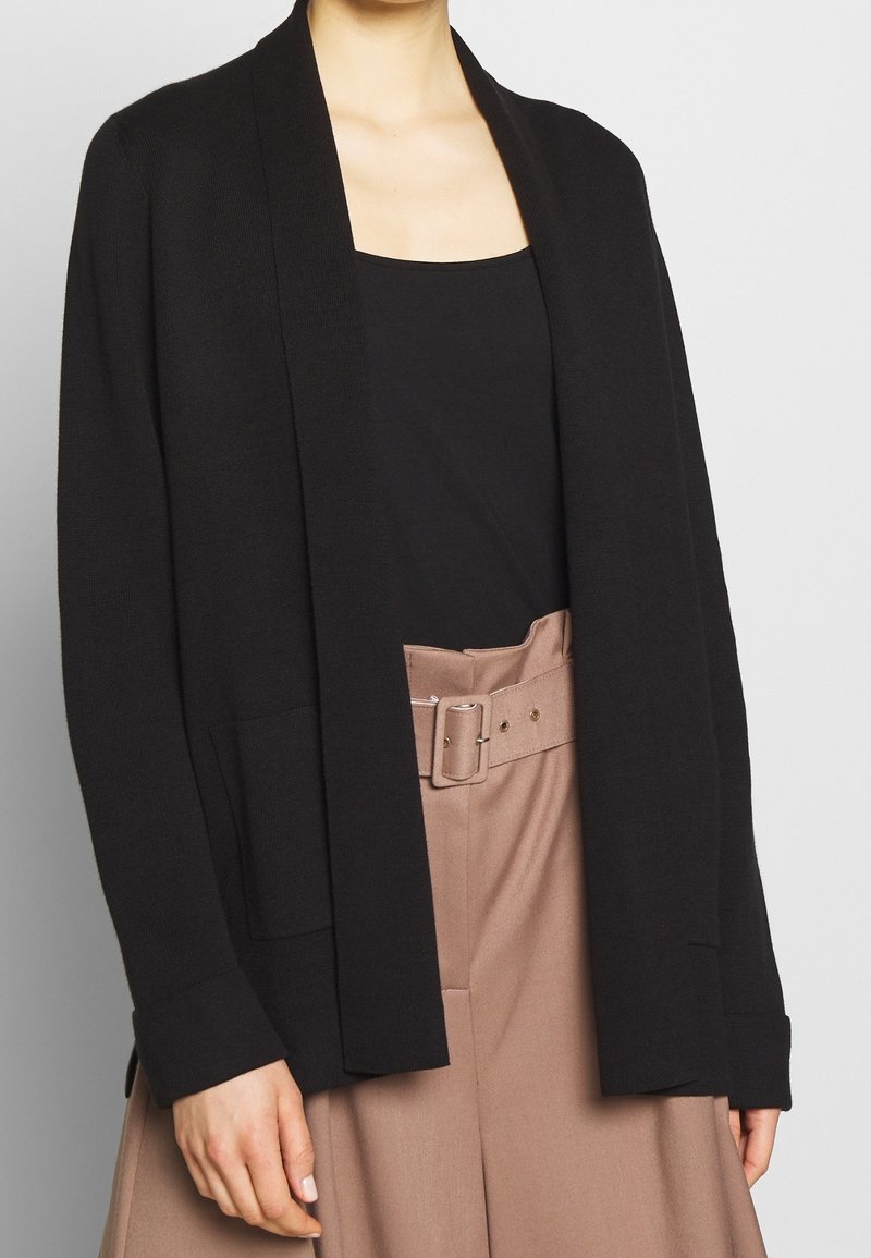 Svart öppen cardigan med avslappnad passform, långa ärmar och sidofickor, bärs över en svart topp och beiga högmidjade byxor.