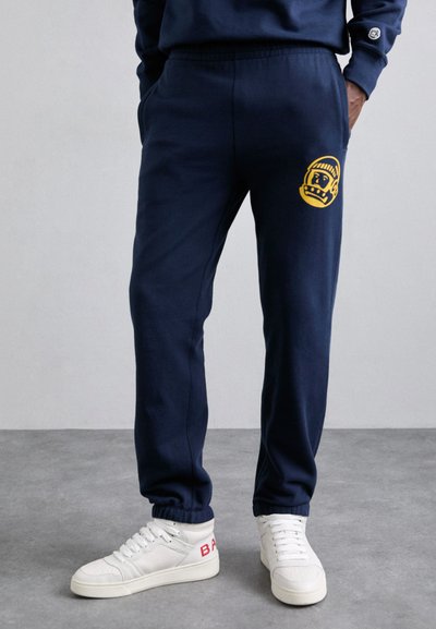 Navy sweatpants med elastisk talje, sidelommer og et gult grafisk motiv på venstre lår, parret med hvide højhælede sneakers.