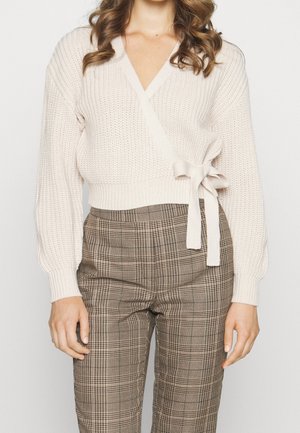 Femme portant un pull cache-cœur en maille crème avec un nœud sur le côté et un pantalon marron à carreaux taille haute.